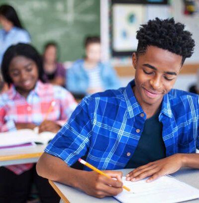 Smiling male student writing in classroom. Teenage boy is concentrating at high school. He is sitting at desk.